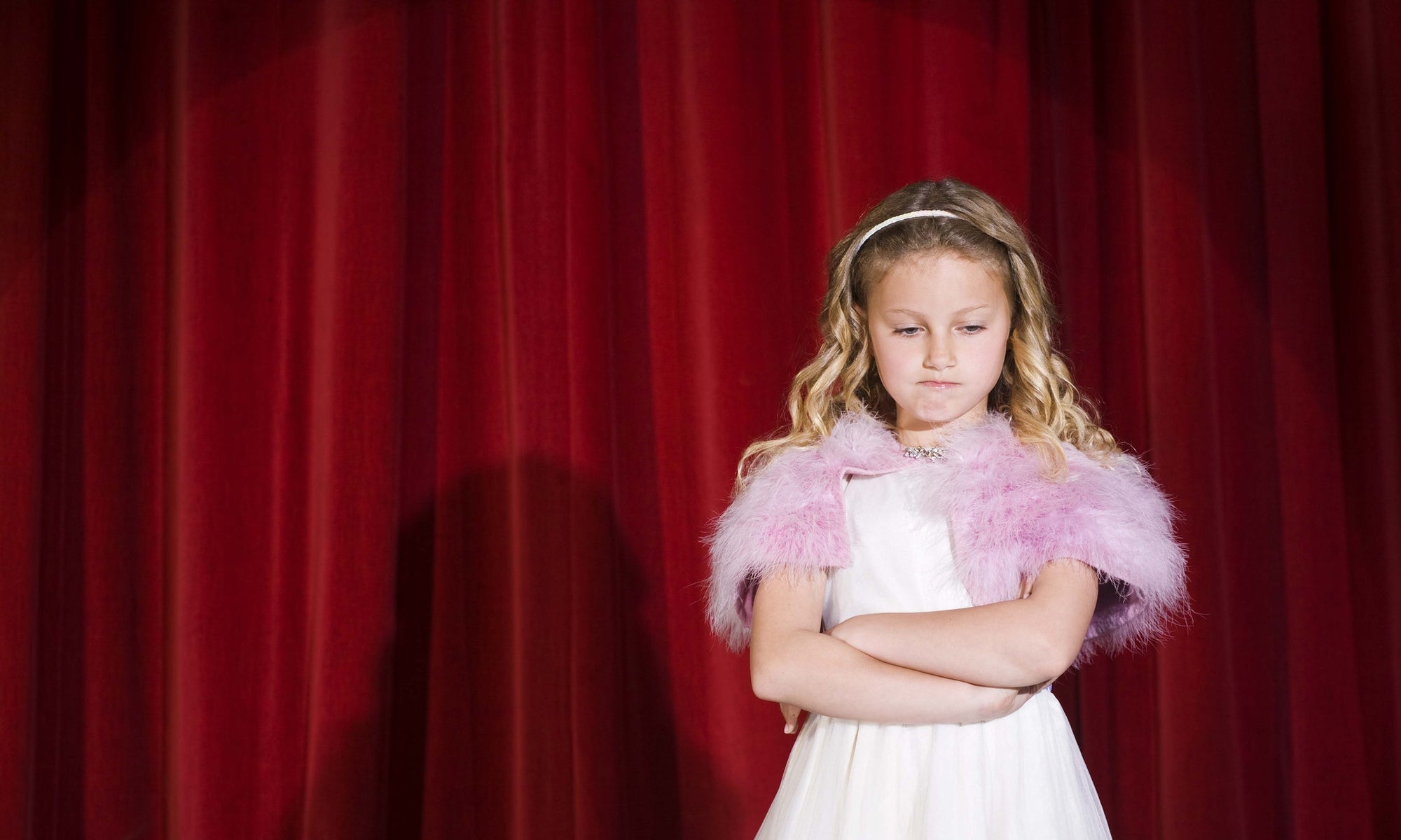 Front view of young girl standing in spotlight against red stage curtain looking annoyed